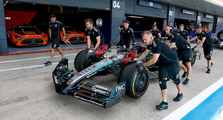 Varios miembros del equipo empujan un carro de carreras de Fórmula 1 Mercedes-AMG PETRONAS fuera del pit lane, con dos carros naranjas Mercedes-AMG GT al fondo.