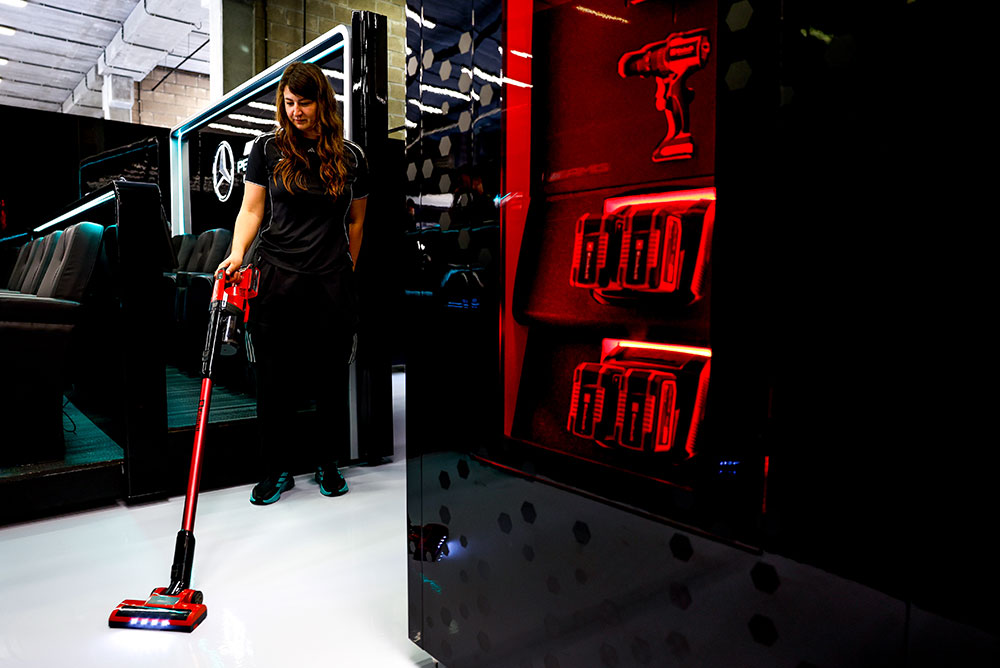 Una integrante del equipo Mercedes-AMG PETRONAS Formula One Team limpia un piso blanco y brillante usando una aspiradora inalámbrica Einhell. A la derecha, unas estanterías iluminadas en rojo exhiben varias baterías Einhell Power X-Change y un taladro inalámbrico.