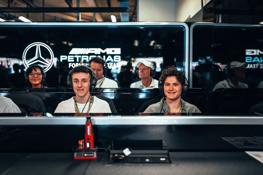 Dos hombres están sentados en el garaje del equipo Mercedes-AMG PETRONAS Formula One, con audífonos puestos, disfrutando la experiencia detrás de escena. En primer plano se aprecia un dispositivo rojo de Einhell.