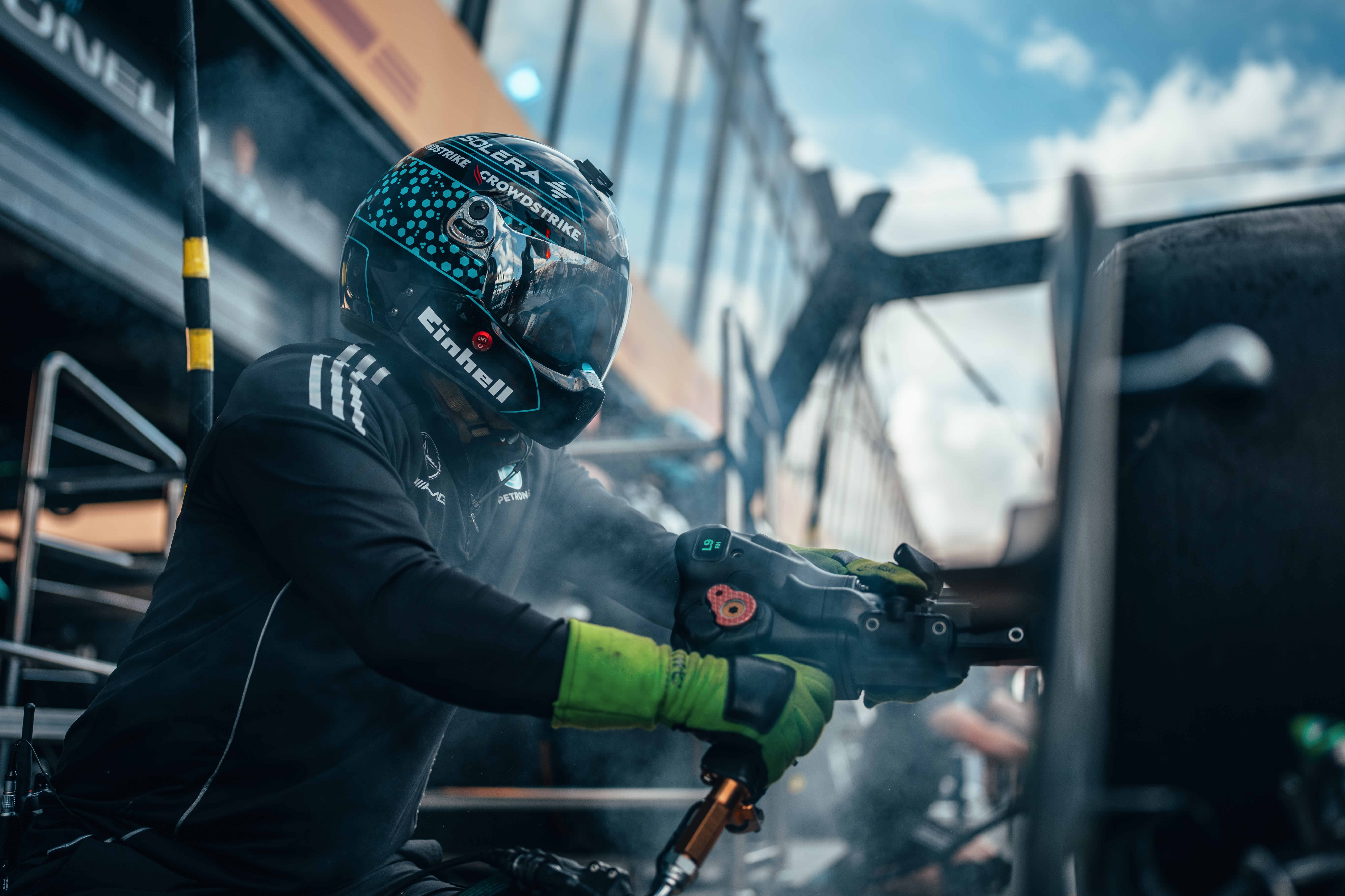 Un integrante del equipo de boxes de Fórmula 1, con casco y guantes, usa una pistola neumática para ruedas durante una parada, rodeado de humo y equipos.