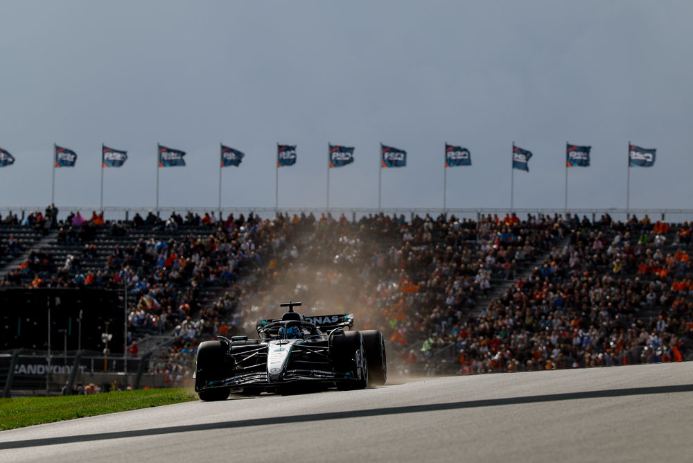 Un auto del equipo Mercedes-AMG PETRONAS Formula One pasa frente a tribunas llenas durante una carrera. Los aficionados agitan banderas al fondo, creando una atmósfera vibrante.
