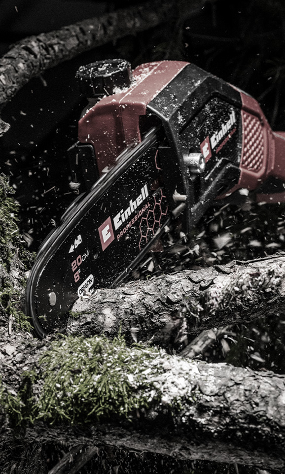 Una motosierra inalámbrica roja de Einhell corta una rama mientras el serrín vuela por el aire.
