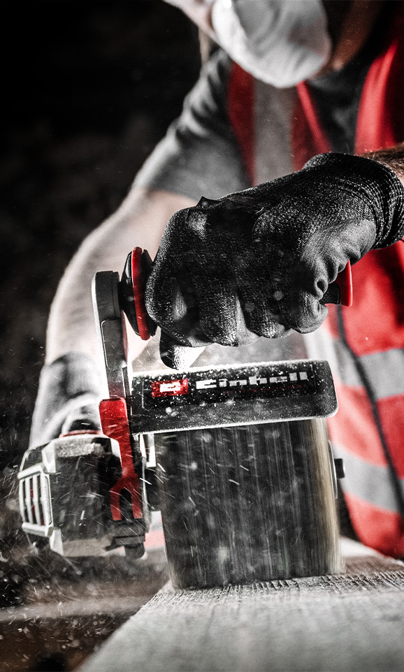 Una persona con guantes negros lija un trozo de madera con una lijadora roja de Einhell, mientras partículas de polvo vuelan por el aire.